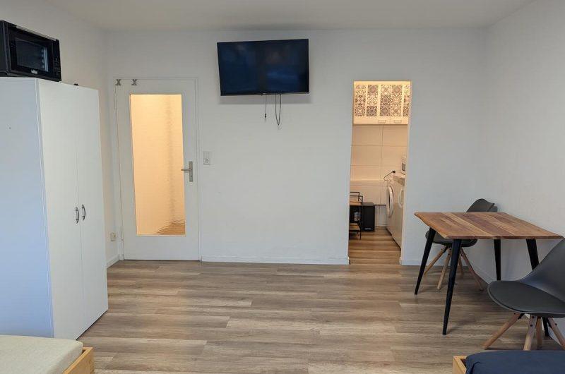 Ansicht des Wohnbereichs mit Wand-TV und Zugang zur Küche.