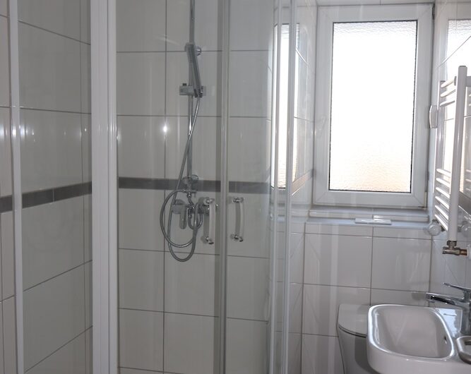 Helles Tageslichtbad mit Glasdusche, Waschmaschinenanschluss und modernen weißen Fliesen.