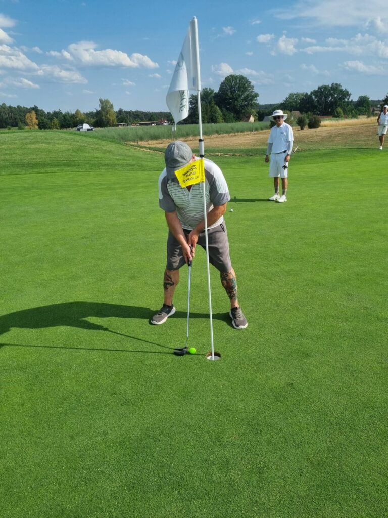 Goldberg Fast Hole in One an Der Bahn 9 | Maderer Immobilien Golf Putting Scene: Man Lining Up a Short Put on a Bright Green Green, Flagstick Nearby, Others Watching in Sunny Course Setting